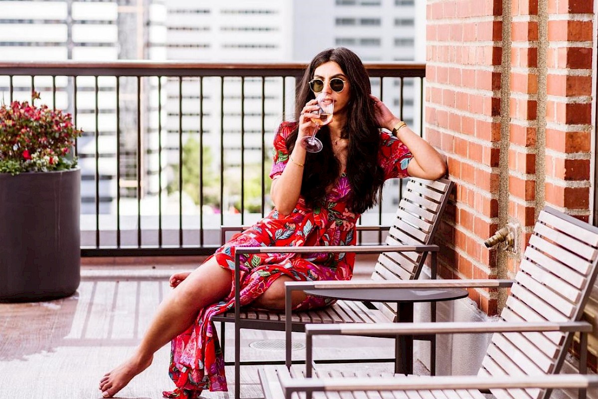 A woman in a red dress with floral patterns sits on a balcony chair, sipping a drink and wearing sunglasses, with a cityscape in the background.