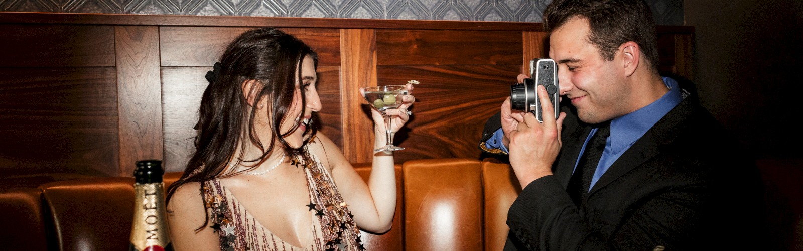 A woman holding a drink poses while a man in a suit takes her photo in a bar or restaurant with a camera.