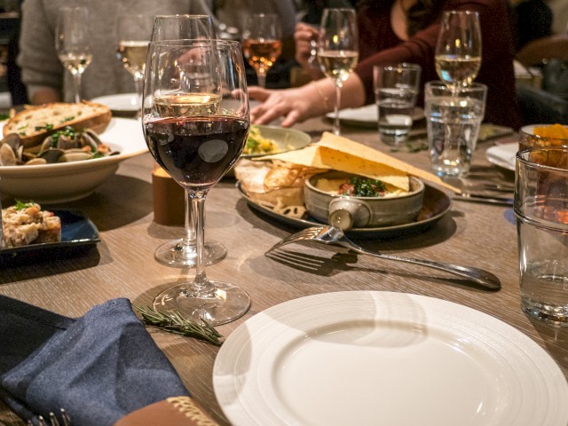 A dining table with various dishes, glasses of wine, water glasses, and cutlery set for multiple people, with hands reaching for food.