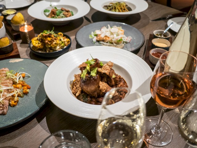 A table set with gourmet dishes, including meats, pasta, and seafood, accompanied by glasses of wine and elegant plating.
