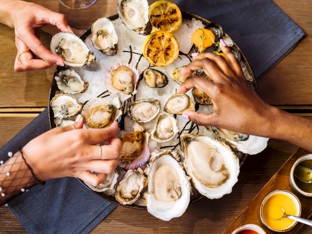 People are enjoying a platter of oysters garnished with lemon halves and sauces on the side, illustrating a communal seafood dining experience.