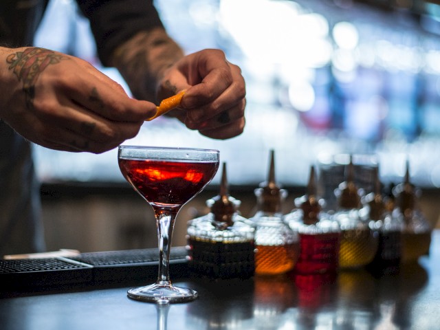 A tattooed person is garnishing a cocktail in a martini glass with an orange peel, with several bottles in the background on the counter.
