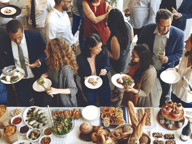 A group of people is gathered around a table filled with an assortment of food, engaging in conversation and enjoying their meals.