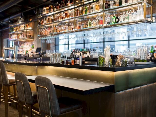 The image shows a modern bar setting with high chairs, a counter lined with bottles, glassware, and bar tools against a backdrop of a wide mirror.