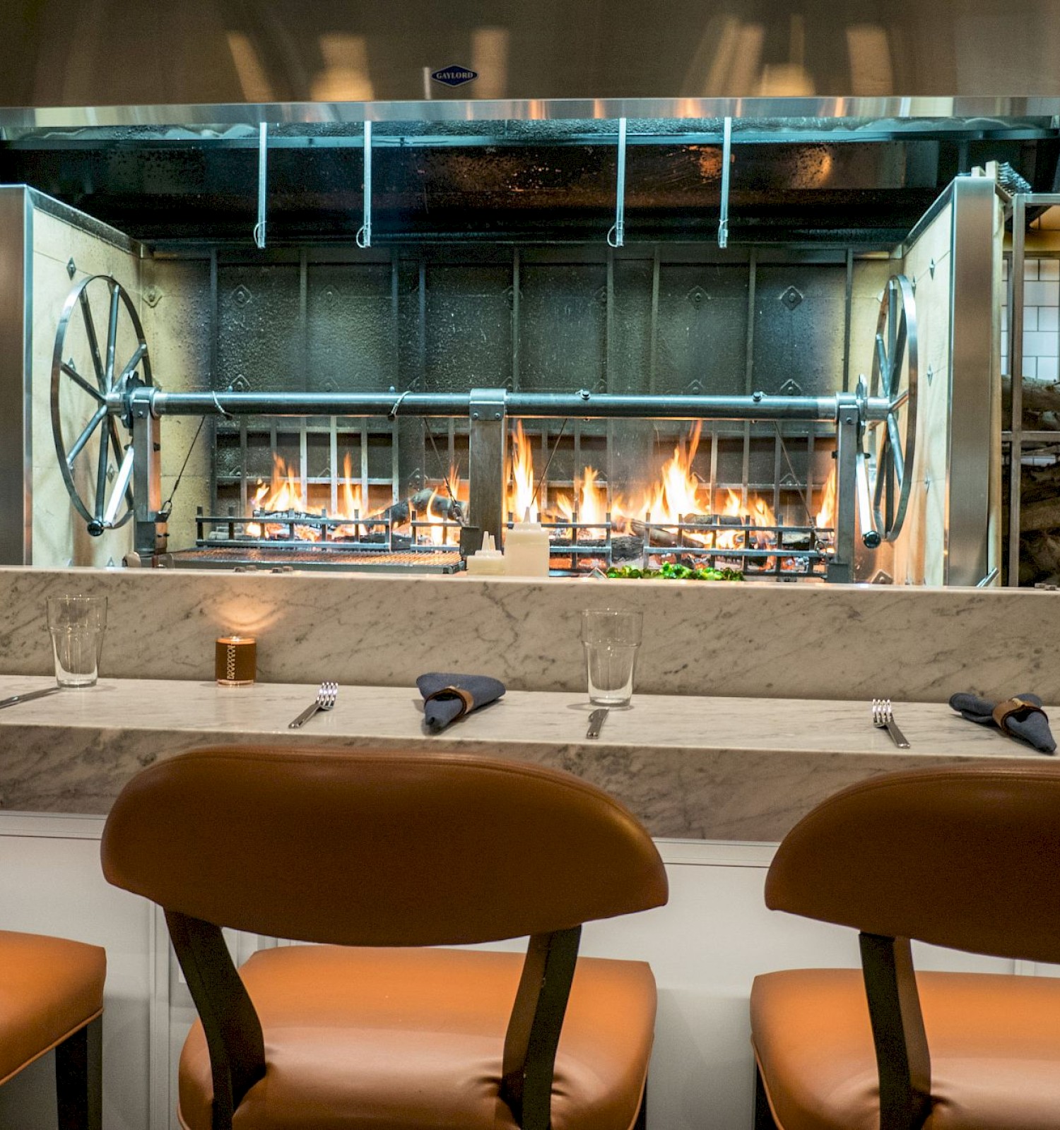 A modern restaurant kitchen with an open flame grill is seen, with seating in front and place settings on the marble countertop.