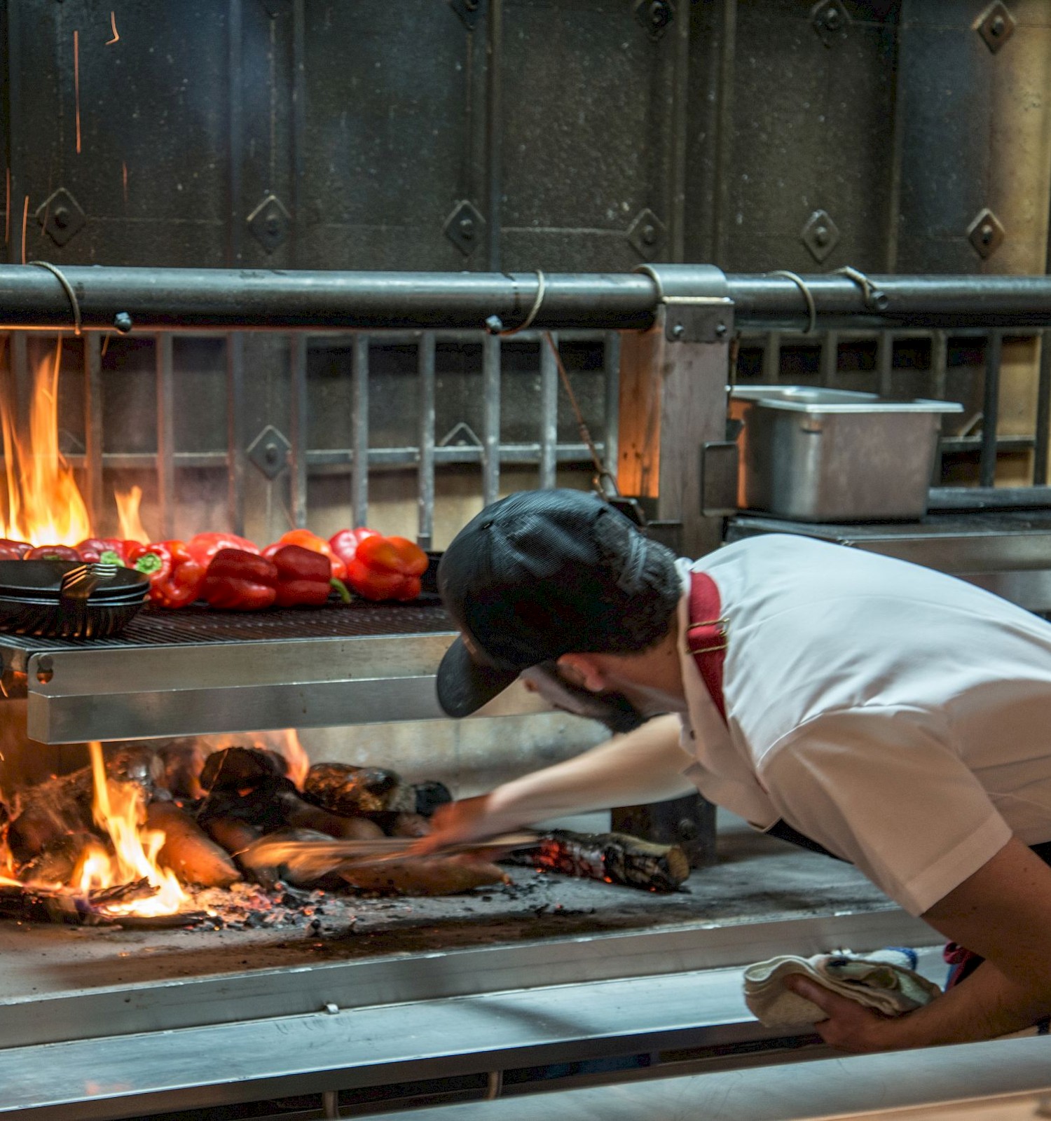 A person is tending to a grill with flames and red peppers, wearing a cap and apron, while adjusting logs or coal in a busy kitchen setting.