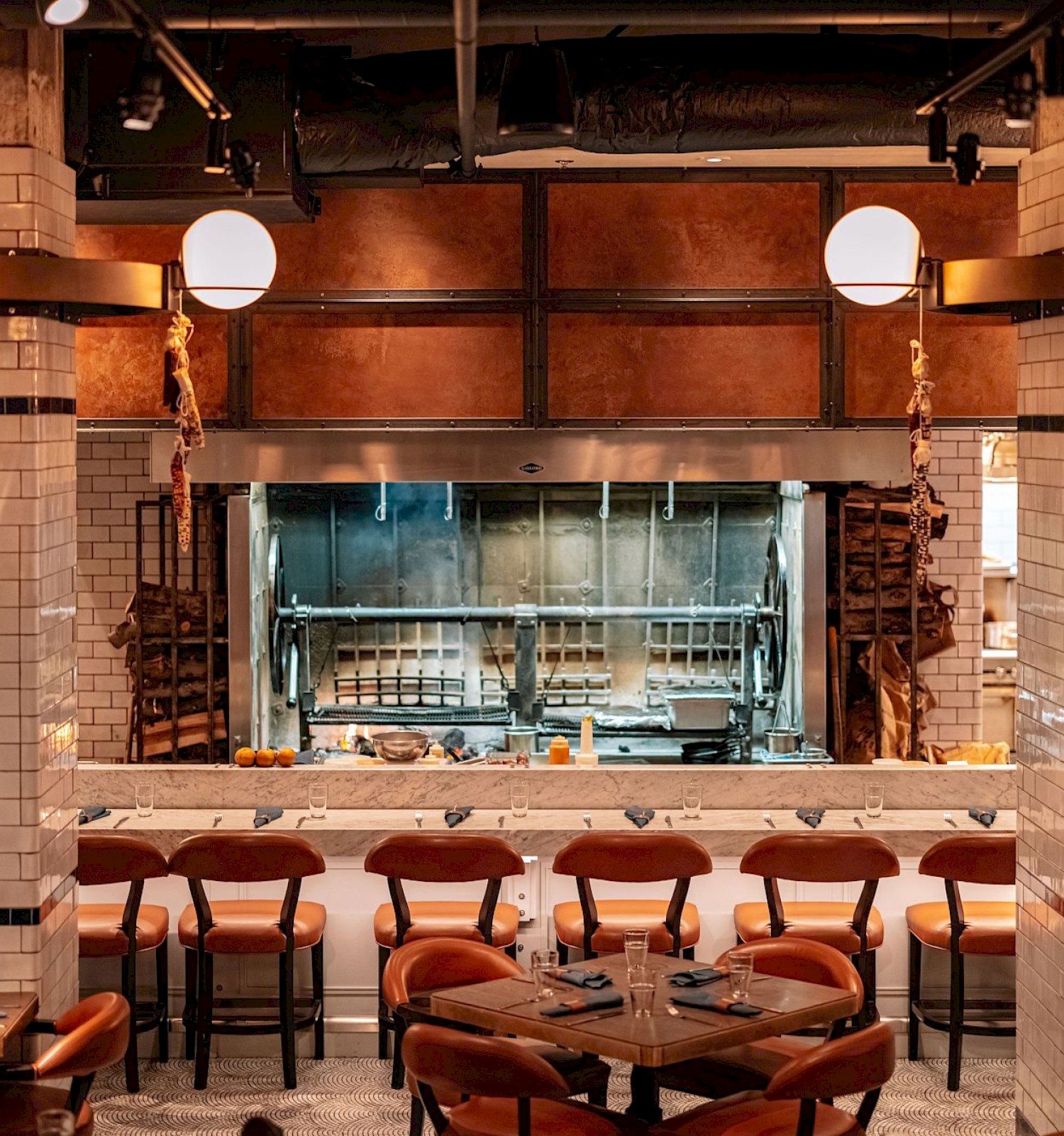 The image shows a cozy restaurant interior with a grill area, tiled walls, warm lighting, and tables set with chairs.