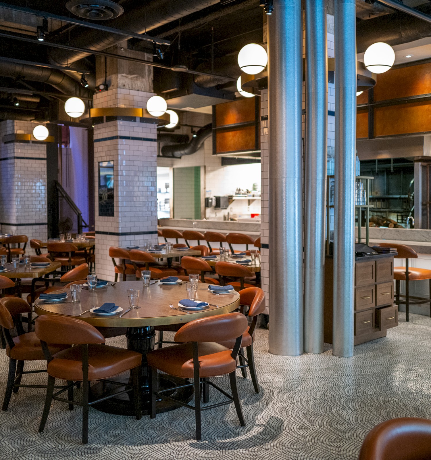 A modern restaurant interior with round tables, leather chairs, industrial lighting, and an open kitchen design.