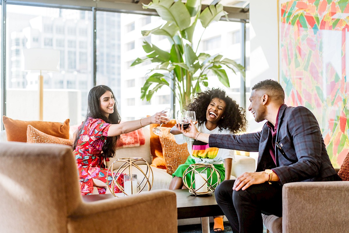 Three people sitting indoors, smiling and toasting with drinks in a brightly decorated and modern setting with large windows and plants.