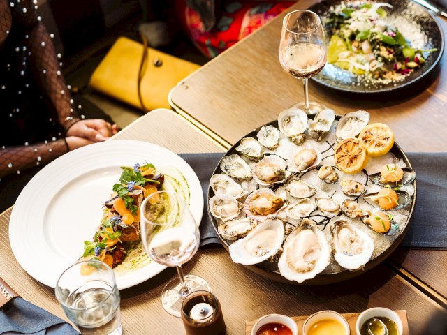The image shows a table with a seafood platter, likely oysters, along with various sauces, a glass of wine, and two other plates of food.
