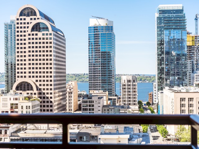The image shows a cityscape with modern skyscrapers, a body of water in the background, and a foreground railing.