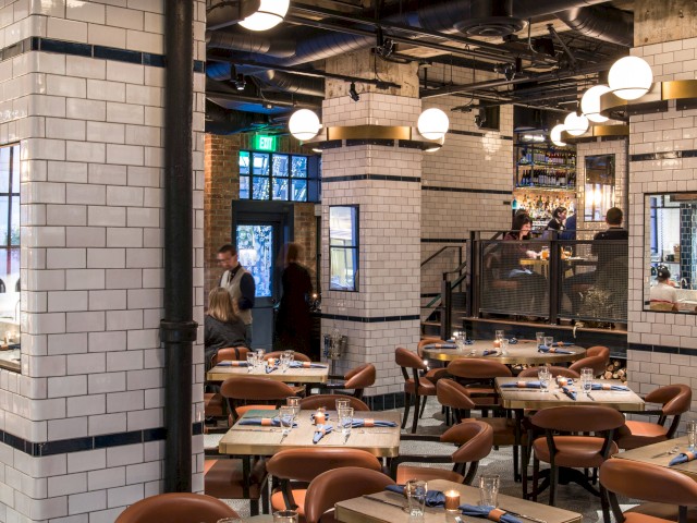 An upscale, modern restaurant with white tiled walls, wooden tables, brown chairs, and round ceiling lights, creating a cozy dining atmosphere.