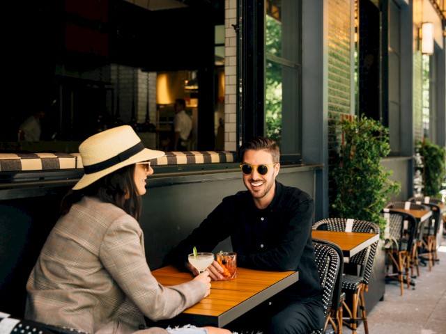 Two people are sitting at an outdoor café, enjoying drinks and talking, with greenery and additional seating visible in the background.