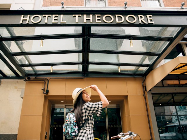 A person in a checkered dress and hat stands with a bicycle in front of Hotel Theodore, smiling and looking upward.