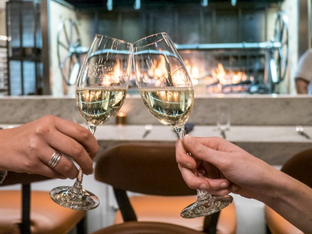 Two people clink wine glasses in a toast, with a cozy, well-lit fireplace in the background, creating a warm and inviting atmosphere.