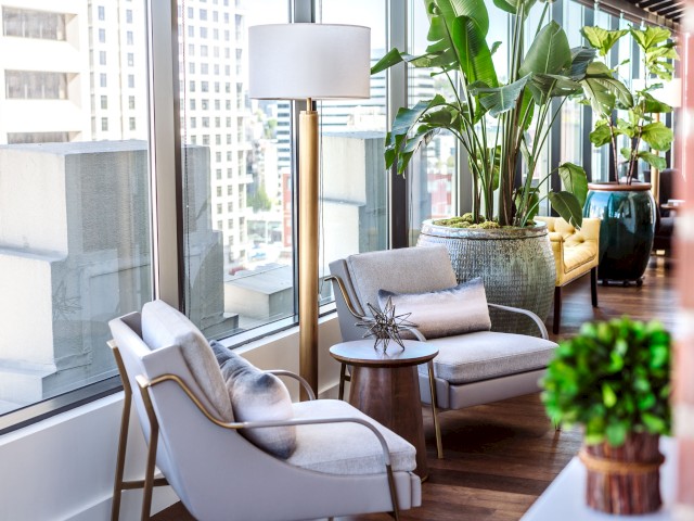 A modern lounge with large windows, two chairs, a small table, potted plants, and a floor lamp, offering a view of an urban cityscape outside.