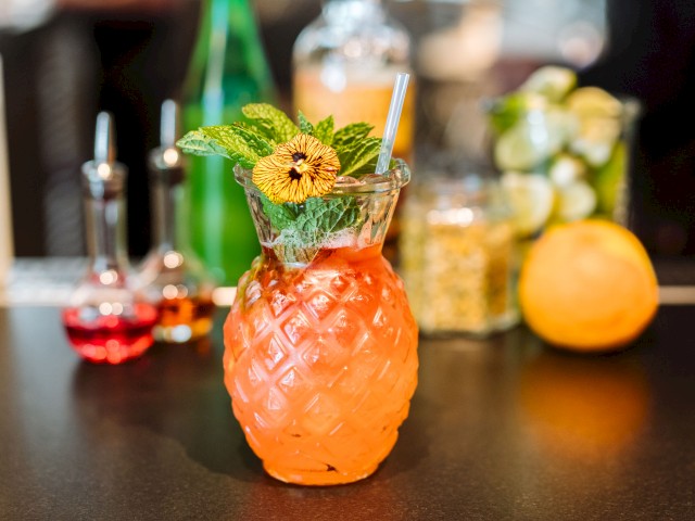 A tropical cocktail served in a pineapple-shaped glass with a straw and garnished with mint and a flower, placed on a bar counter.
