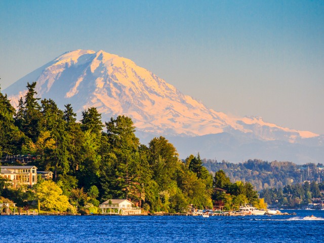 The image showcases a scenic view of a large snow-capped mountain with a lush green forest and houses near a body of water in the foreground.
