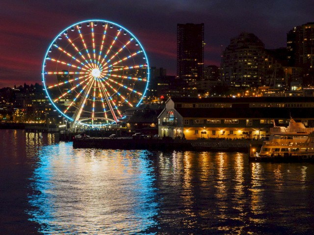 A brightly lit Ferris wheel by the waterfront at night, with city buildings and lights reflecting on the water’s surface.