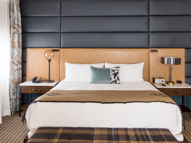 A neatly made bed with a headboard, two bedside tables with lamps, a phone, and a curtain-covered window in a modern, cozy hotel room.
