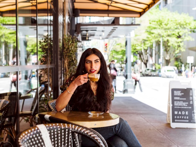 A woman is sitting at an outdoor café, sipping coffee. The setting is urban with trees and a sign in the background indicating 