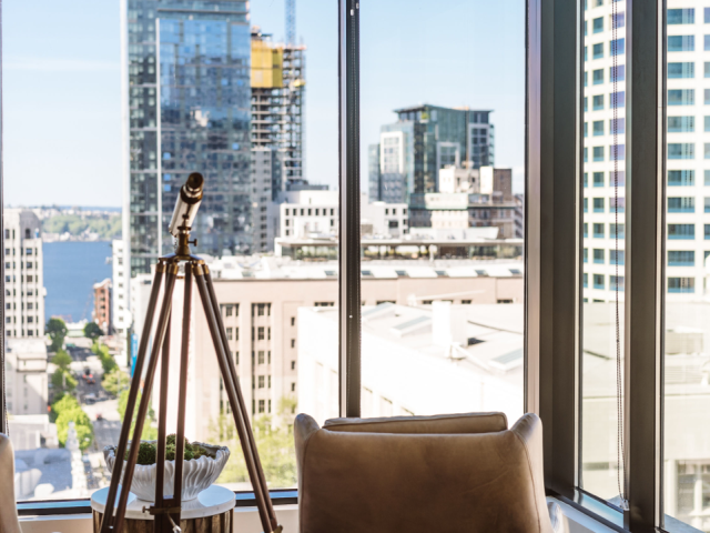 The image shows a modern urban view through large windows, featuring skyscrapers and two armchairs with a telescope on a stand between them.