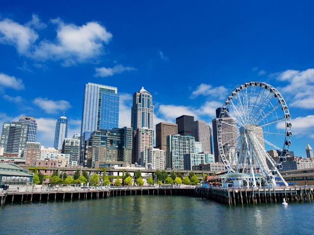 The image shows a waterfront skyline with tall buildings, a Ferris wheel, and a dock area under a blue sky with clouds.