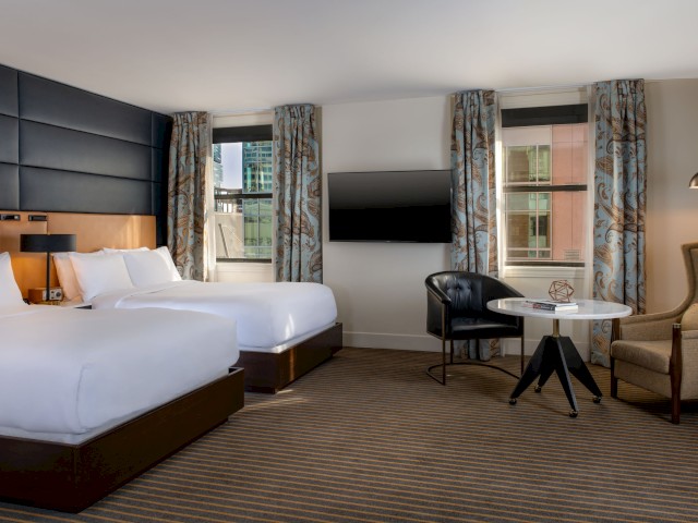 A modern hotel room with two beds, a TV, a round table with chairs, an armchair, and large windows with city views and patterned curtains.