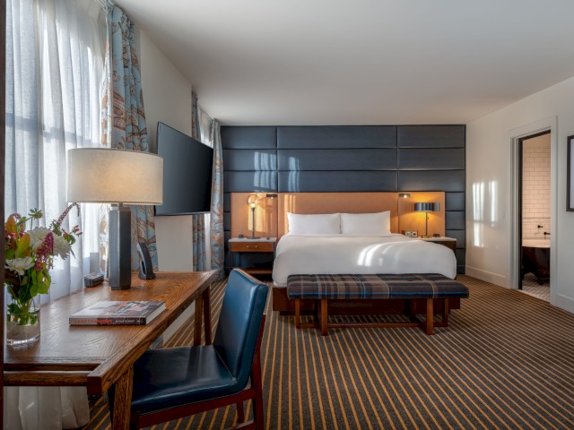 A stylishly decorated hotel room with a king-size bed, desk, TV, and a seating area. A bathroom is visible through an open door.