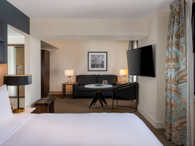 The image shows a modern hotel room with a bed, sofa, coffee table, TV, lamps, and framed artwork on the wall.