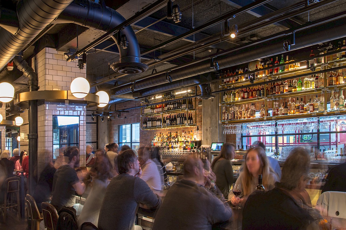 A lively bar with people seated at the counter, shelves stocked with bottles, under warm lighting and industrial decor.