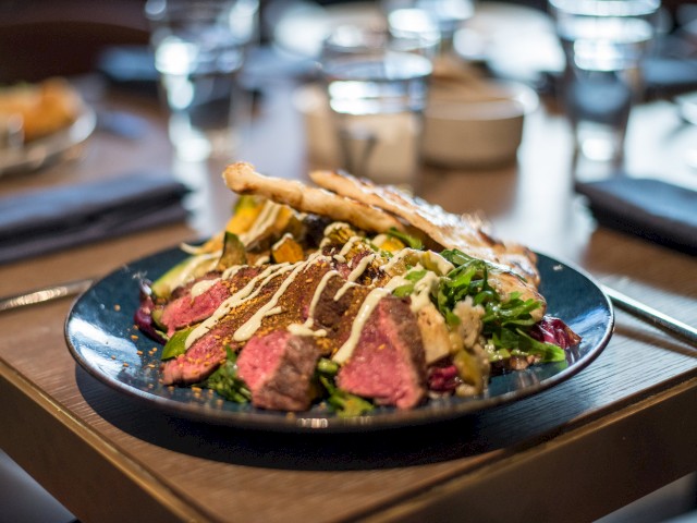 A dish with sliced steak on a bed of greens, topped with sauce, and accompanied by grilled vegetables and flatbread on a dark plate.