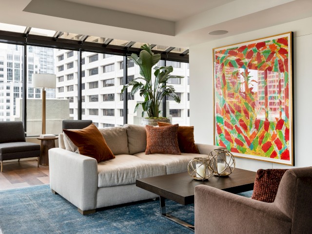 A modern living room with a gray sofa, colorful abstract painting, large windows, plants, and a blue rug completes the scene.