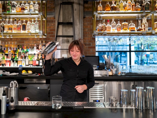 A bartender is shaking a cocktail shaker behind a bar with shelves of liquor bottles and a window view in the background.