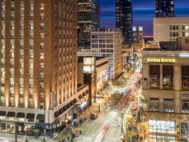 A vibrant cityscape at night with bustling streets, illuminated buildings, and blurred lights from moving traffic on the roads.