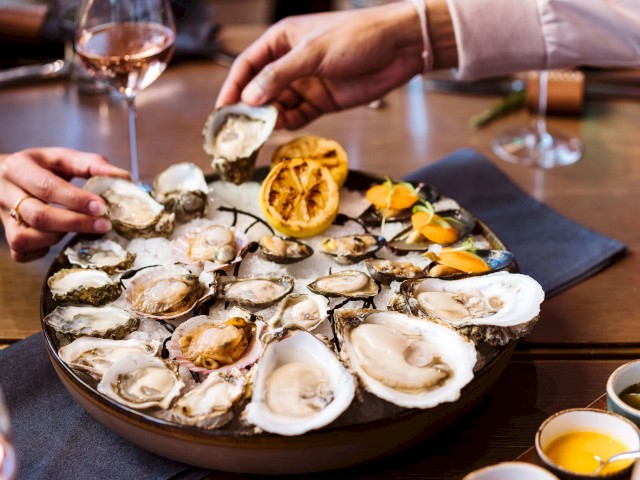 A platter of oysters with lemon and garnishes; people are reaching for them. Wine glasses are in the background.
