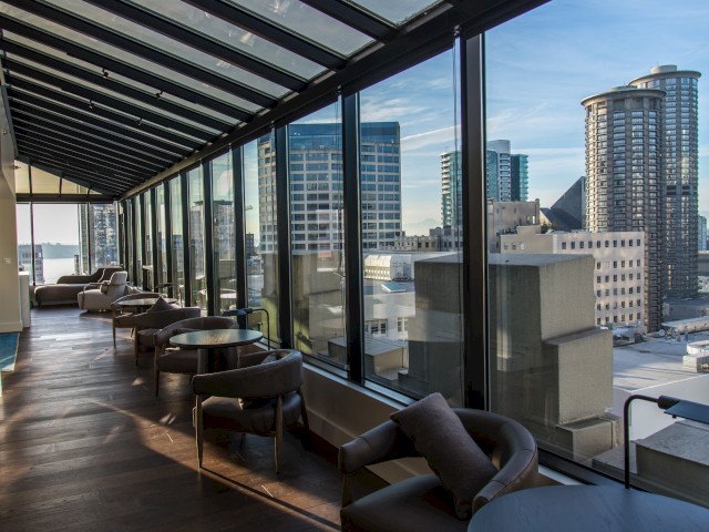 A modern indoor lounge with glass walls offers a cityscape view featuring tall buildings under a clear blue sky.