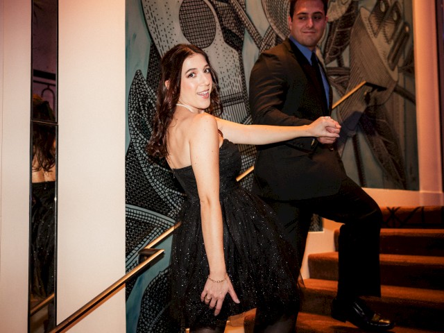 A woman in a black dress and a man in a suit are posing on stairs, with a patterned wall in the background.