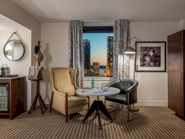 A cozy room with two chairs, a table, mirror, minibar, and wall art, overlooking city buildings through a window.