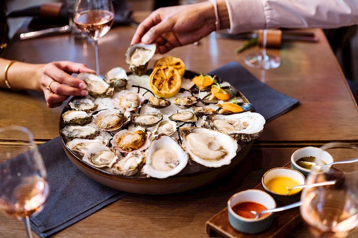 A group enjoys oysters on a platter with lemon, accompanied by sauces and glasses of rosé wine on a dining table.