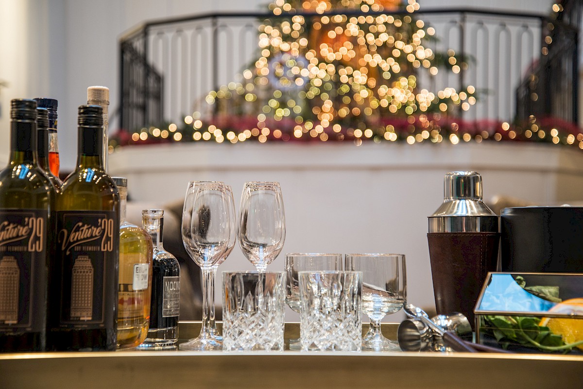 The image shows a bar setup with bottles, glasses, and a cocktail shaker, blurred lights in the background, indicating a festive setting.