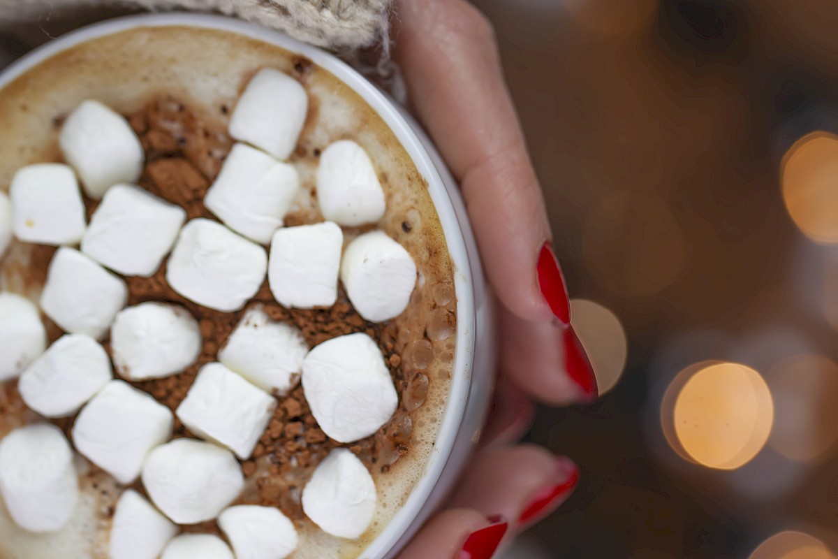 A person holds a cup of hot chocolate topped with marshmallows. They wear a knitted sleeve, with blurred lights in the background.