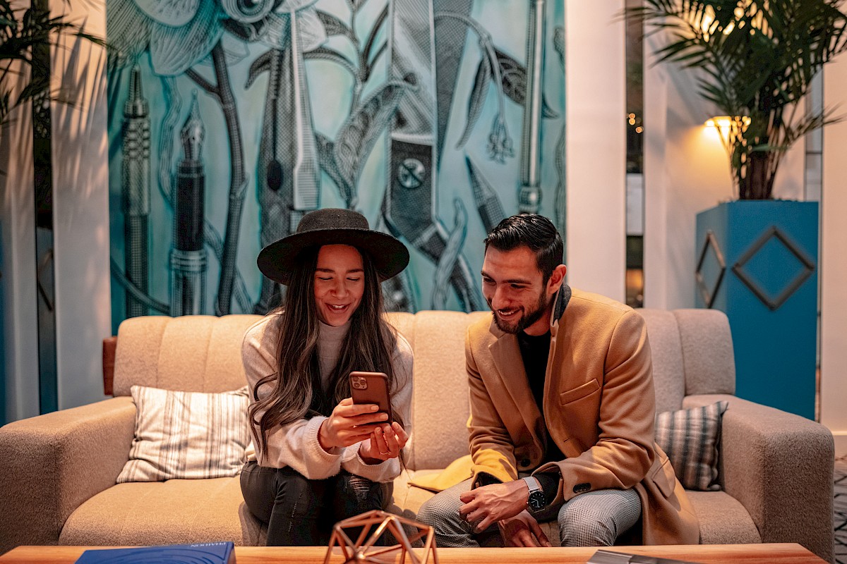 Two people sit on a couch, looking at a phone, with a colorful mural in the background and plants around them.