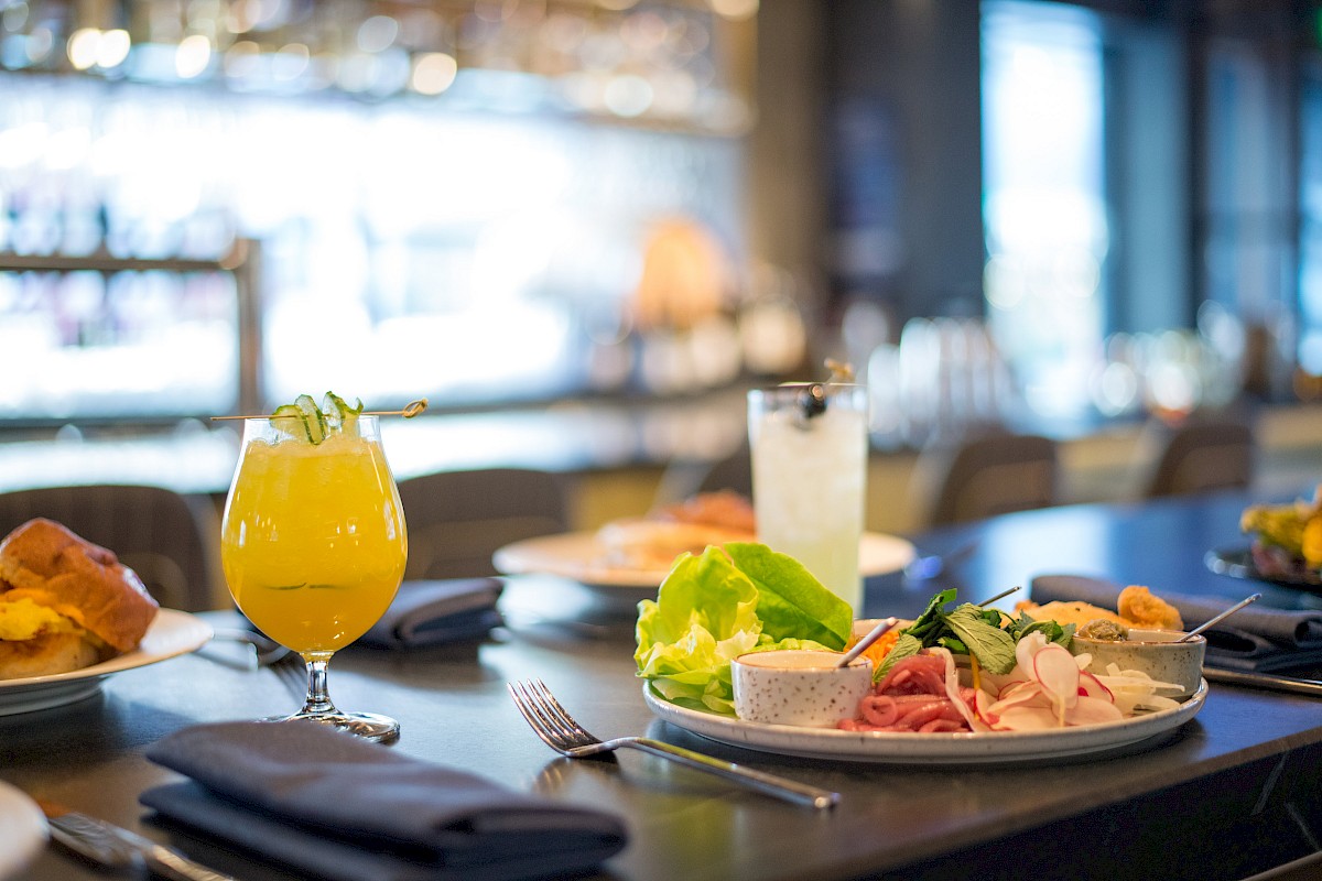 A dining table with a salad, sliced meats, beverages, and bread is set in a restaurant with a blurred background.