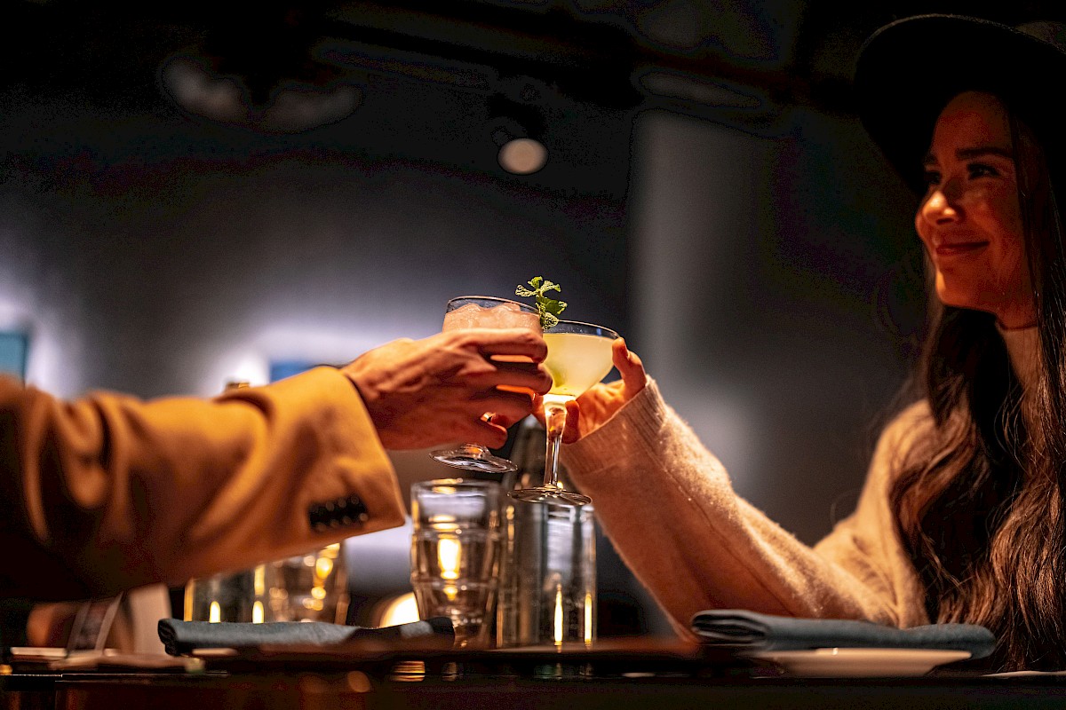 Two people are toasting with cocktails at a dimly lit restaurant.