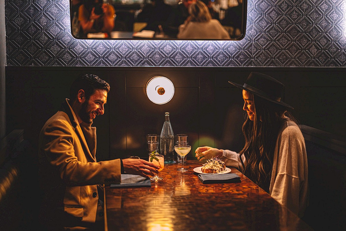 A man and woman are seated in a dimly lit restaurant booth, engaged in conversation with drinks and food on the table.