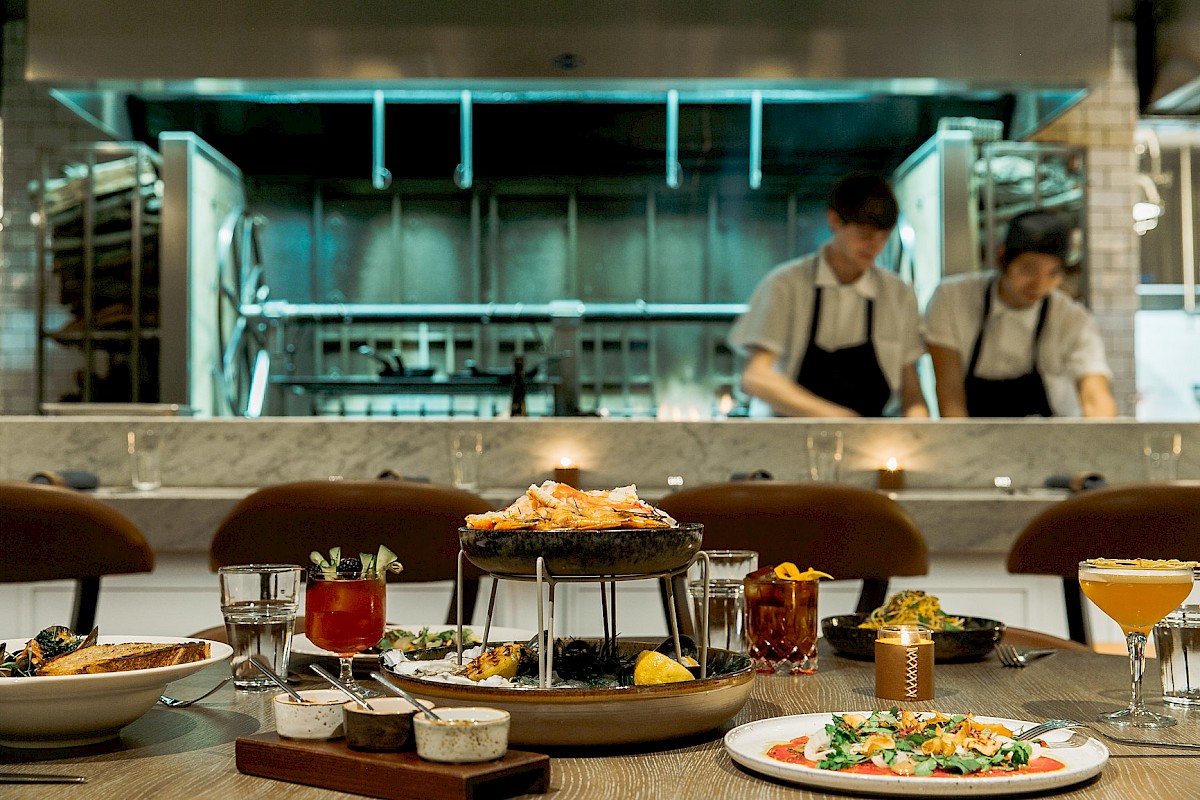 A restaurant setting with a spread of dishes and drinks on a table, and chefs working in the kitchen in the background.