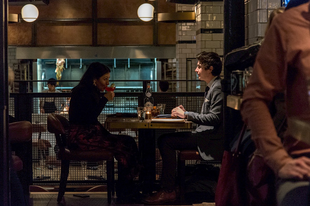 A man and woman are dining at a restaurant, sitting across from each other at a table, with a kitchen visible in the background.