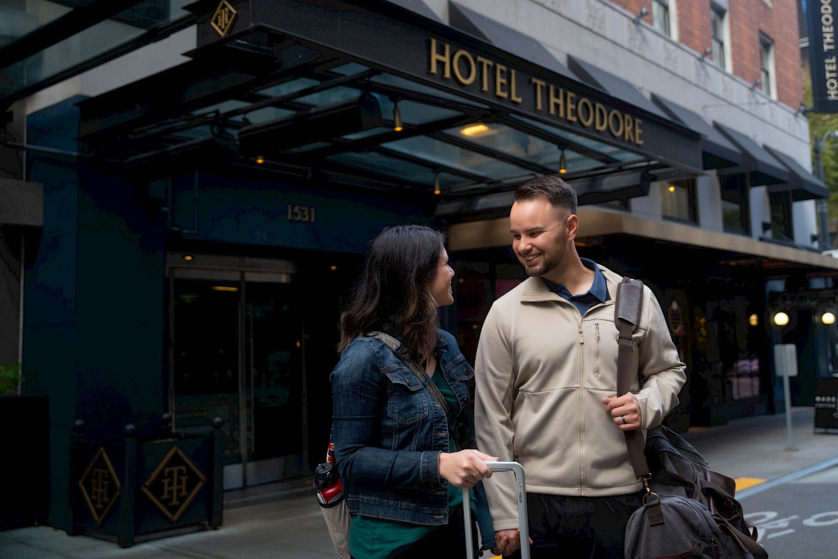 A man and woman with luggage stand outside Hotel Theodore, smiling at each other on the sidewalk.