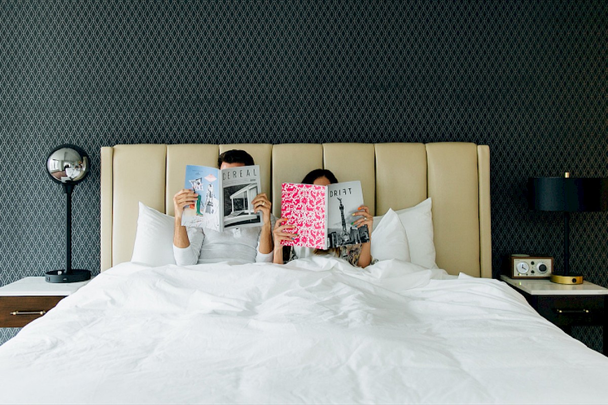 Two people are sitting up in bed, each reading a magazine, with a modern bedside setup and a textured wall behind them.